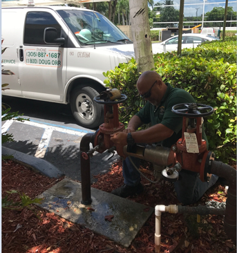 A person kneels on the ground installing a backflow preventer with a white Douglas Orr Plumbing truck parked in the background
